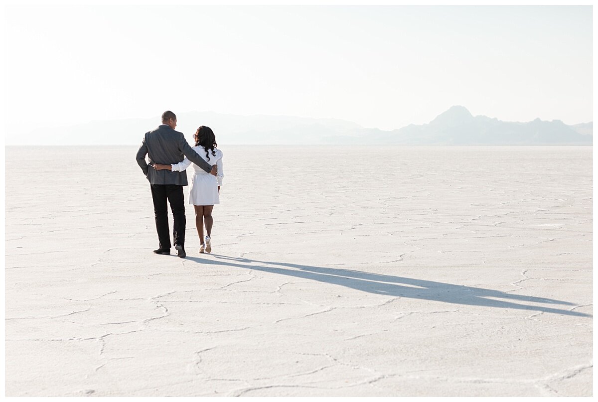 Couple walks with hands around each other’s waists across the Bonneville Salt Flats. Blue mountains out in the horizon capture the beauty of this epic location for engagement pictures.