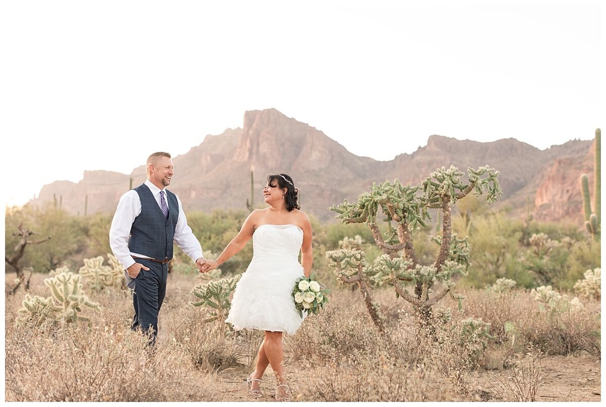Bride and Groom on their wedding day walking through the desert hand in hand. Surrounded by towering saguaro cacti and mountains in the background.