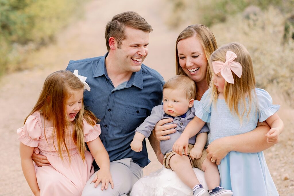 Family Photos in the desert as family snuggles close. Mom holds youngest infant son on her lap while toddler sisters lean on Mom and Dad's legs.