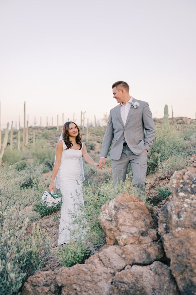 Bride and groom walking up a rocky hillside surrounded by saguaros, sharing a loving look on their wedding day in the Arizona desert