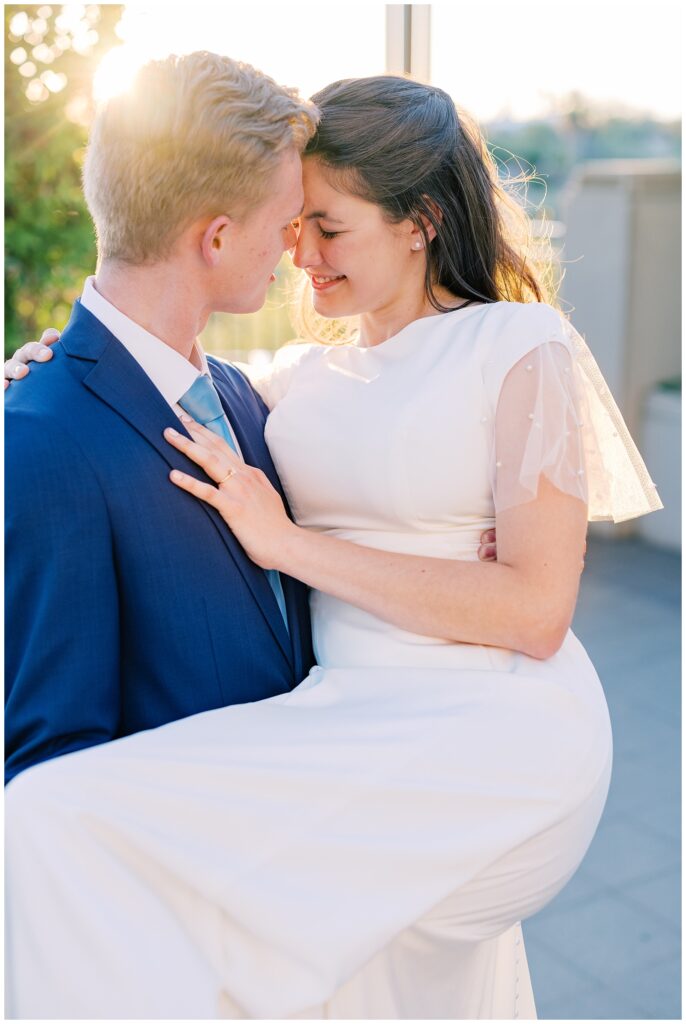 Groom holds bride on their wedding day. They lean in close while the desert sun peeks in behind them.